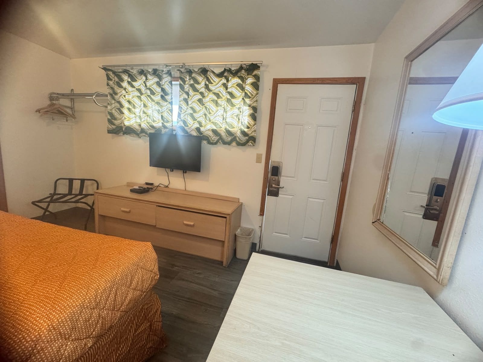 A room with a bed, a desk, a television, a door, and a window with patterned curtains.