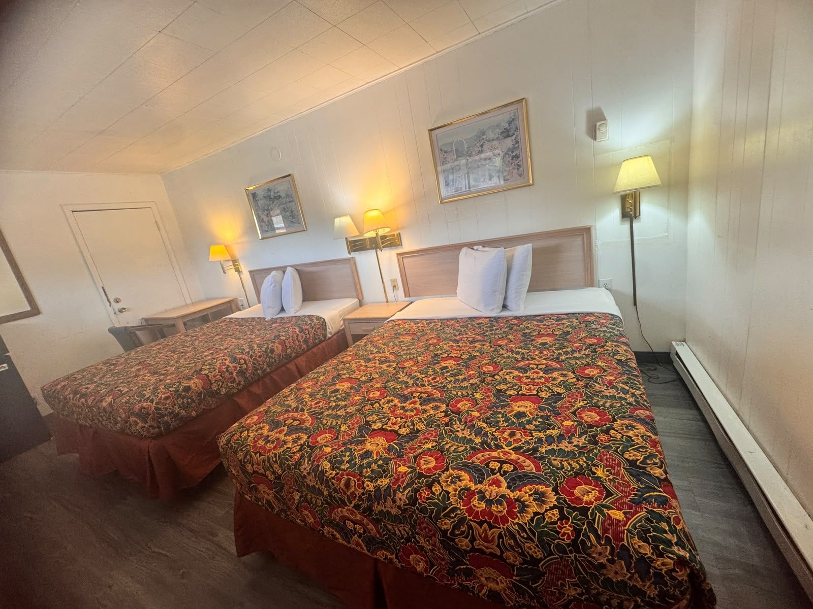 Una camera d'albergo con due letti, ognuno con una coperta a motivi floreali, una testiera in legno e lampade ai comodini.