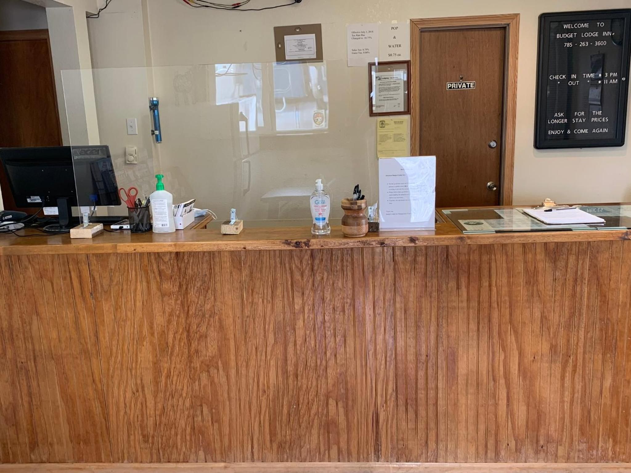 A reception desk with a wooden finish, a computer monitor, a bottle of hand sanitizer, a water bottle, and a clipboard with papers.