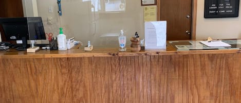 A reception desk with a wooden finish, a computer monitor, a bottle of hand sanitizer, a water bottle, and a clipboard with papers.