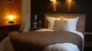 Una cama bien hecha con una manta marrón y almohadas blancas, dos lámparas fijadas a la pared y una mesita de noche con una lámpara y un jarrón de flores.