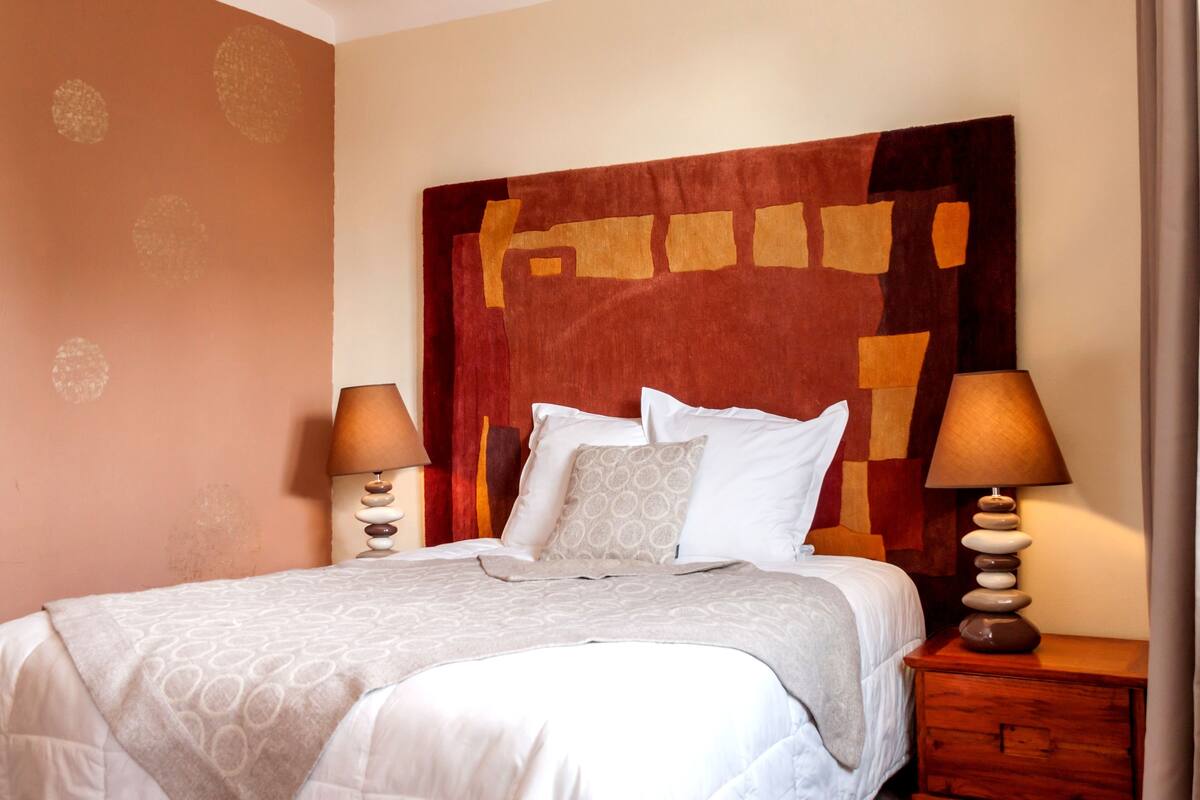 A neatly made bed with a patterned bedspread, two bedside lamps, and a wooden nightstand with a stack of stones.