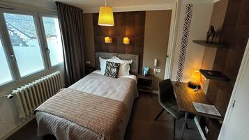 A hotel room with a bed, bedside tables, a desk with a chair, a window, and a radiator.