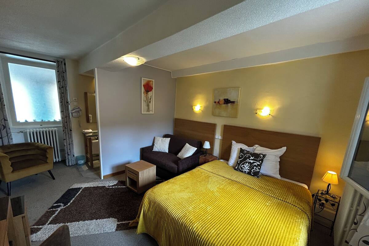 A hotel room with a bed, a sofa, a small table, a chair, a radiator, a window with curtains, and wall-mounted lights.