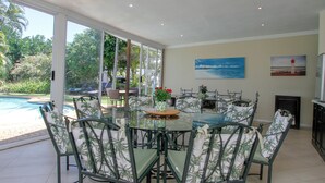 A dining area with a glass table and chairs, a pool visible through large windows, and a painting of a lighthouse on the wall. - THE SANDRINGHAM B&B AND SELF-CATERING (Umhlanga)
