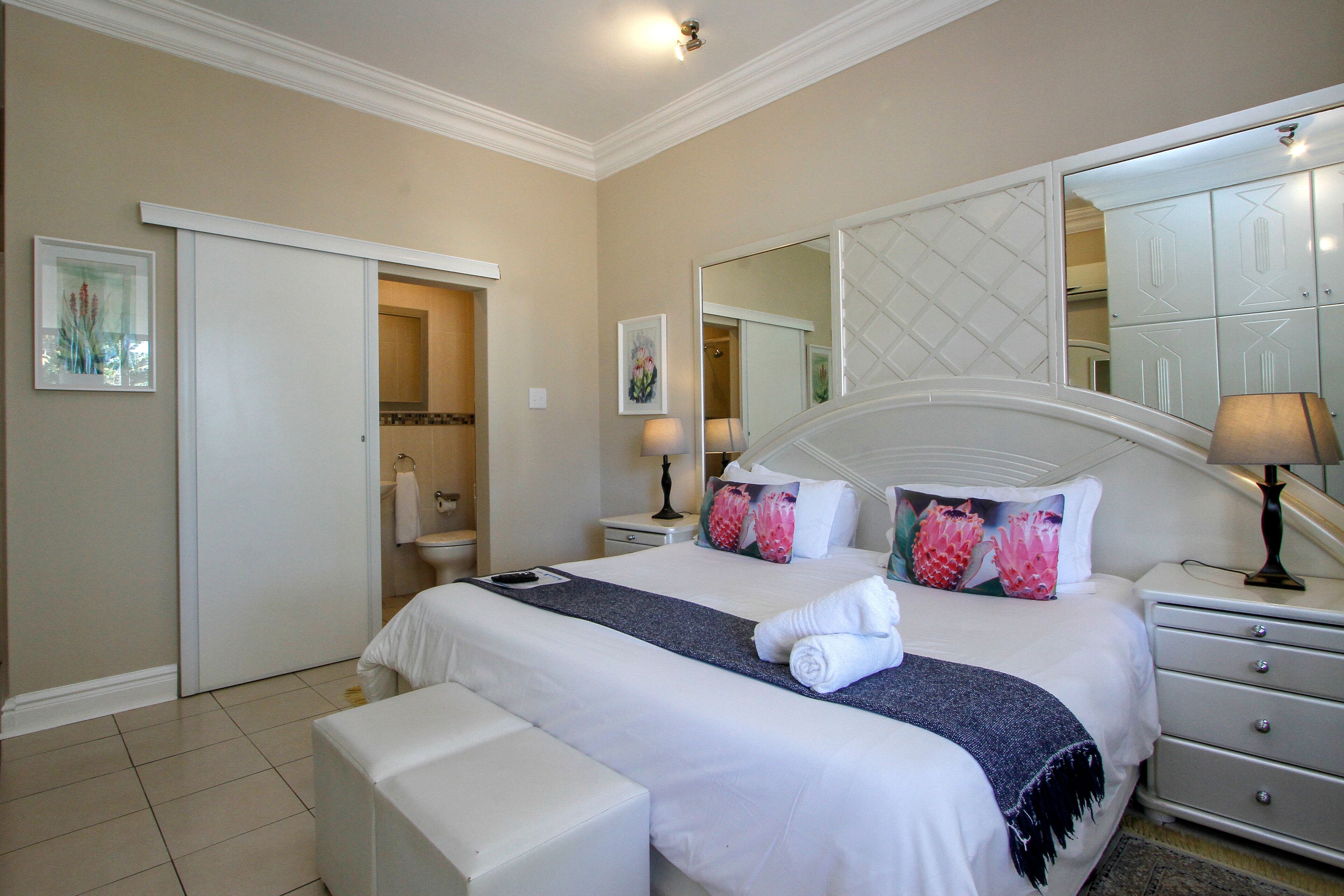A hotel room with a large bed, a bedside table, a nightstand, a bench, a bathroom with a toilet and sink, and a mirror.