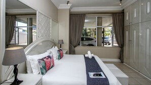 A hotel room with a bed, a nightstand, a lamp, a window with curtains, and a view of the street. - THE SANDRINGHAM B&B AND SELF-CATERING (Umhlanga)