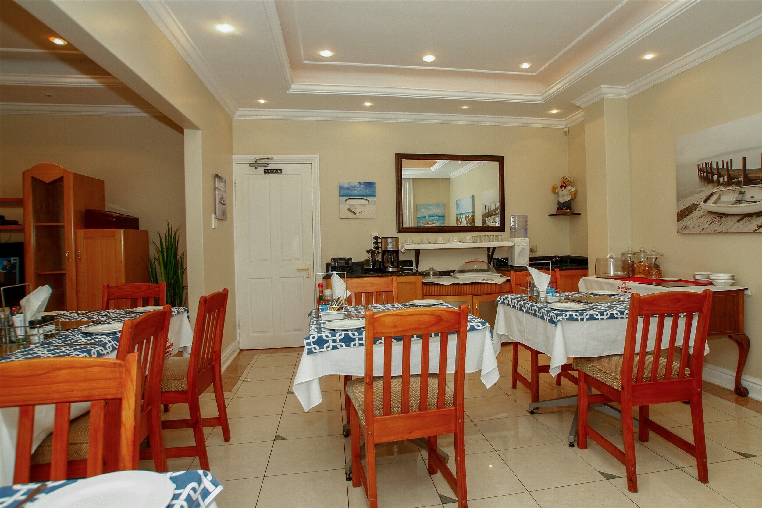 A dining area with wooden tables and chairs, a kitchen in the background, and a mirror on the wall.