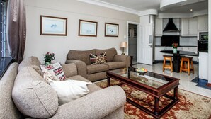 A living room with a sofa, a coffee table, and a rug. There is a kitchen visible in the background. - THE SANDRINGHAM B&B AND SELF-CATERING (Umhlanga)