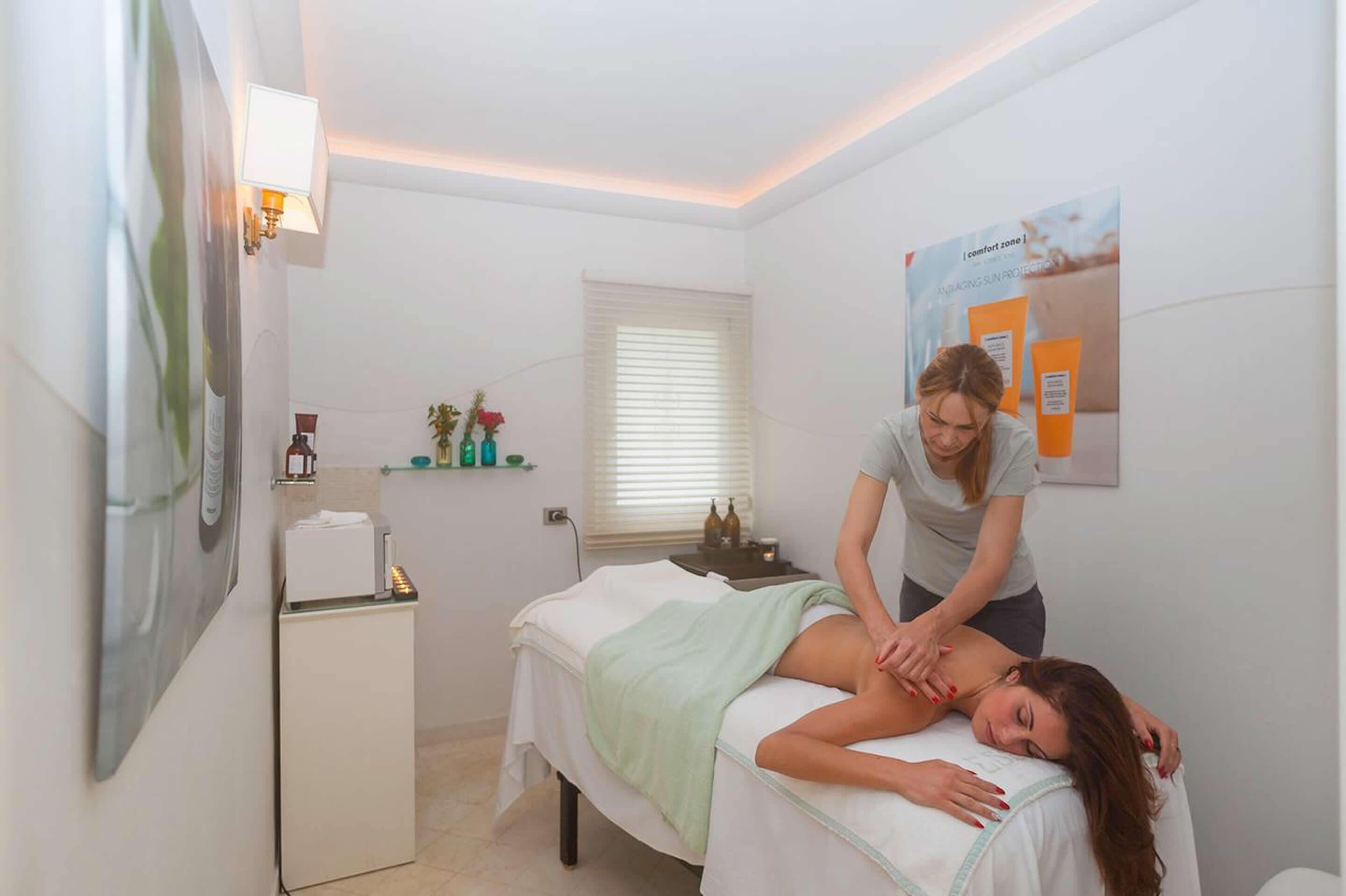 Une salle de massage équipée d’une table de massage, d’un petit meuble, d’une fenêtre avec des stores et d’une affiche accrochée au mur.