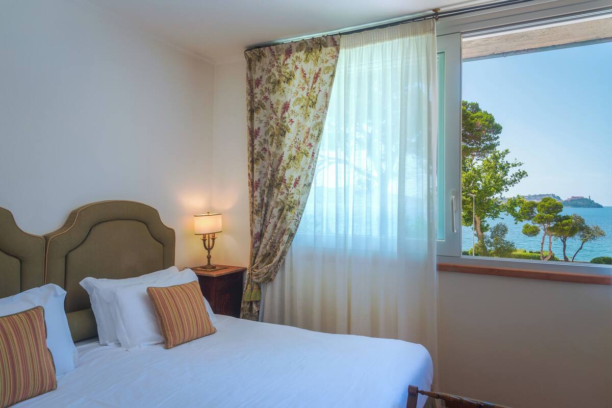 Habitación de hotel con cama, mesita de noche, lámpara y vista al mar a través de una ventana.