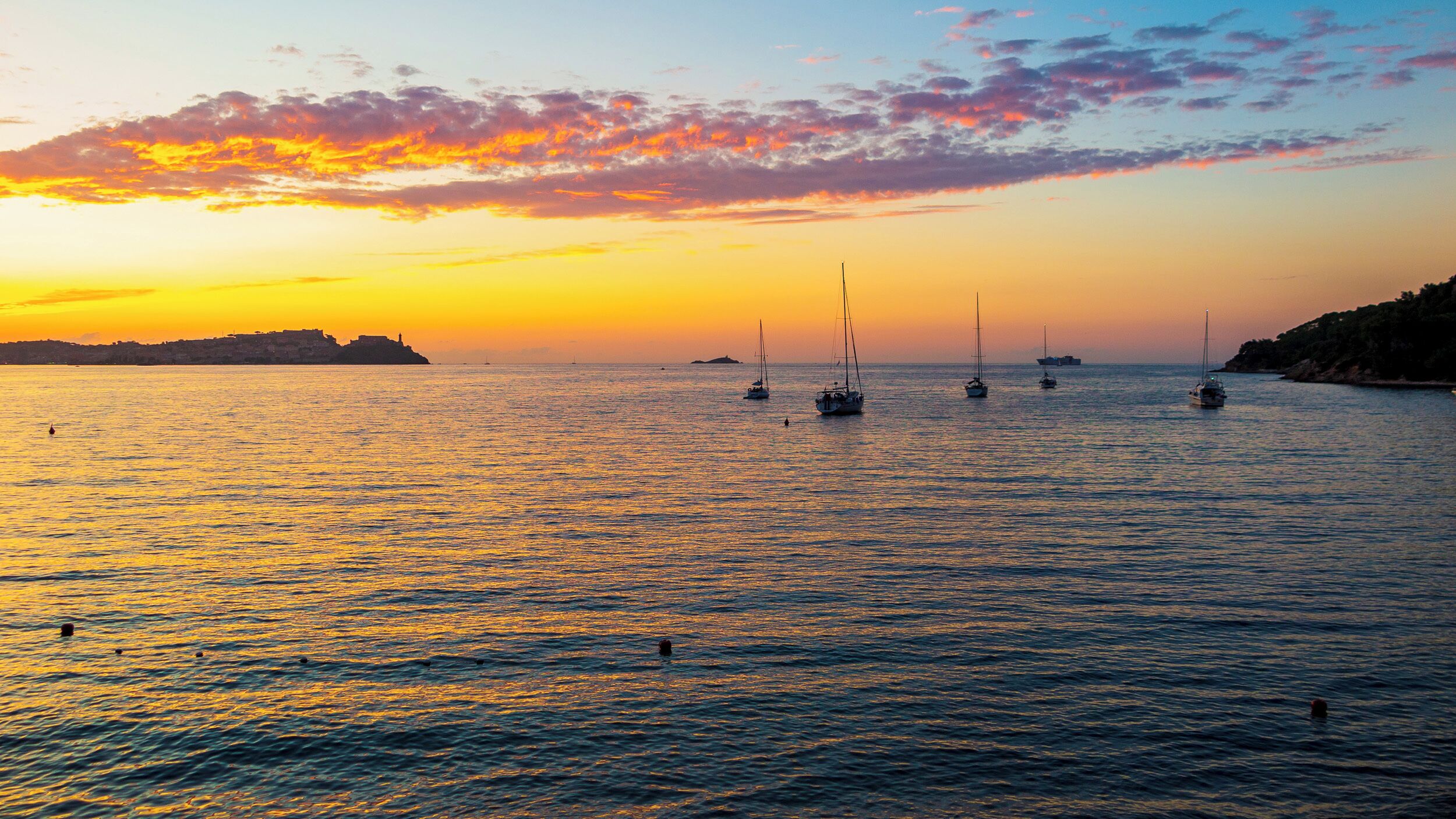 A sunset over a calm sea with several sailboats.