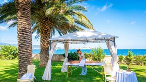 Ein Massagesetup mit Baldachin, Massagetisch und zwei Stühlen in einer grünen Wiese mit Palmen und Blick aufs Meer.
