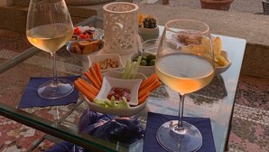 Ein Tisch ist gedeckt mit zwei Gläsern Weißwein, einer Zierkerze und verschiedenen Snacks, darunter Selleriestangen und Nüsse, mit Blick auf einen Sonnenuntergang über dem Meer.
