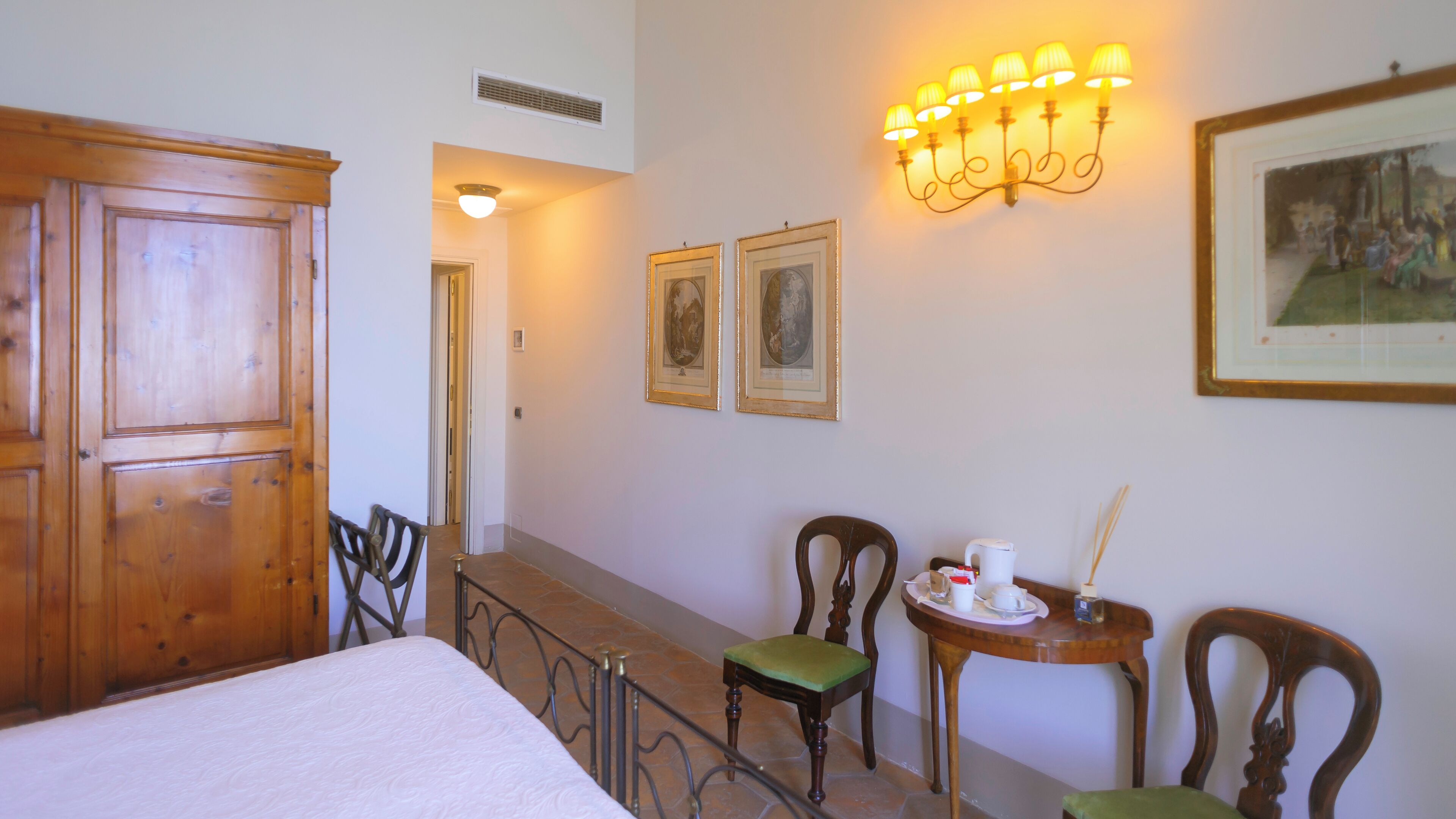 Habitación de hotel con armario de madera, una cama, dos sillas, una mesita con bandeja e iluminación en la pared.