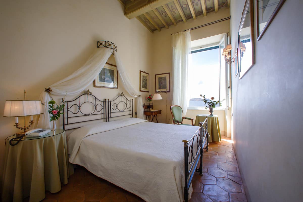 Un dormitorio ordenado con cama, mesitas de noche, una silla y una ventana con cortinas.