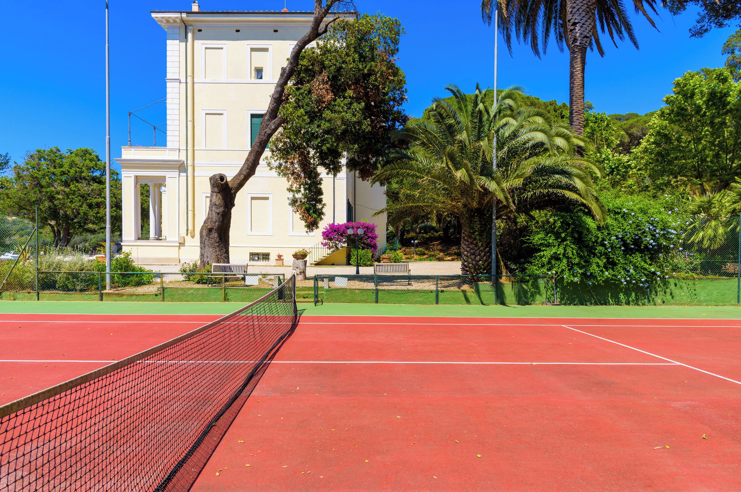 A tennis court with a net, surrounded by greenery and a building with columns.
