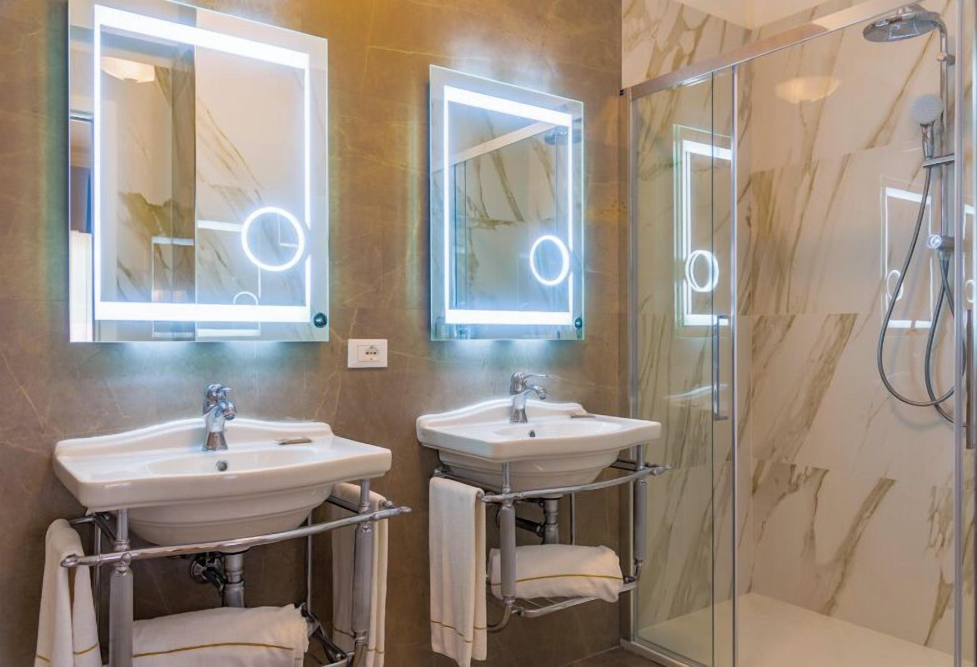 A bathroom with two sinks, a glass shower enclosure, and two illuminated mirrors.