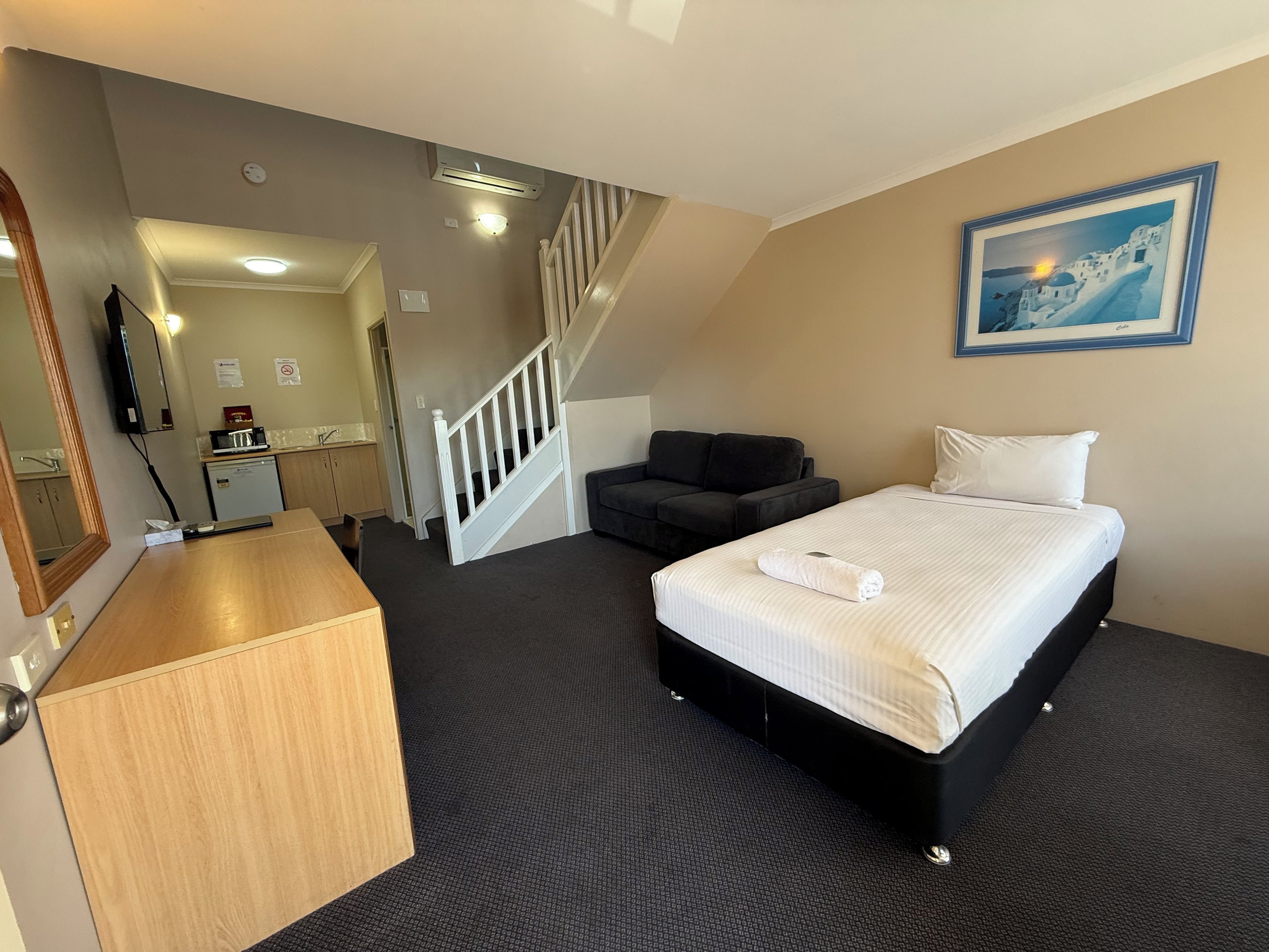 Habitación de hotel con cama, escritorio, silla, sofá, escalera y baño visible al fondo.