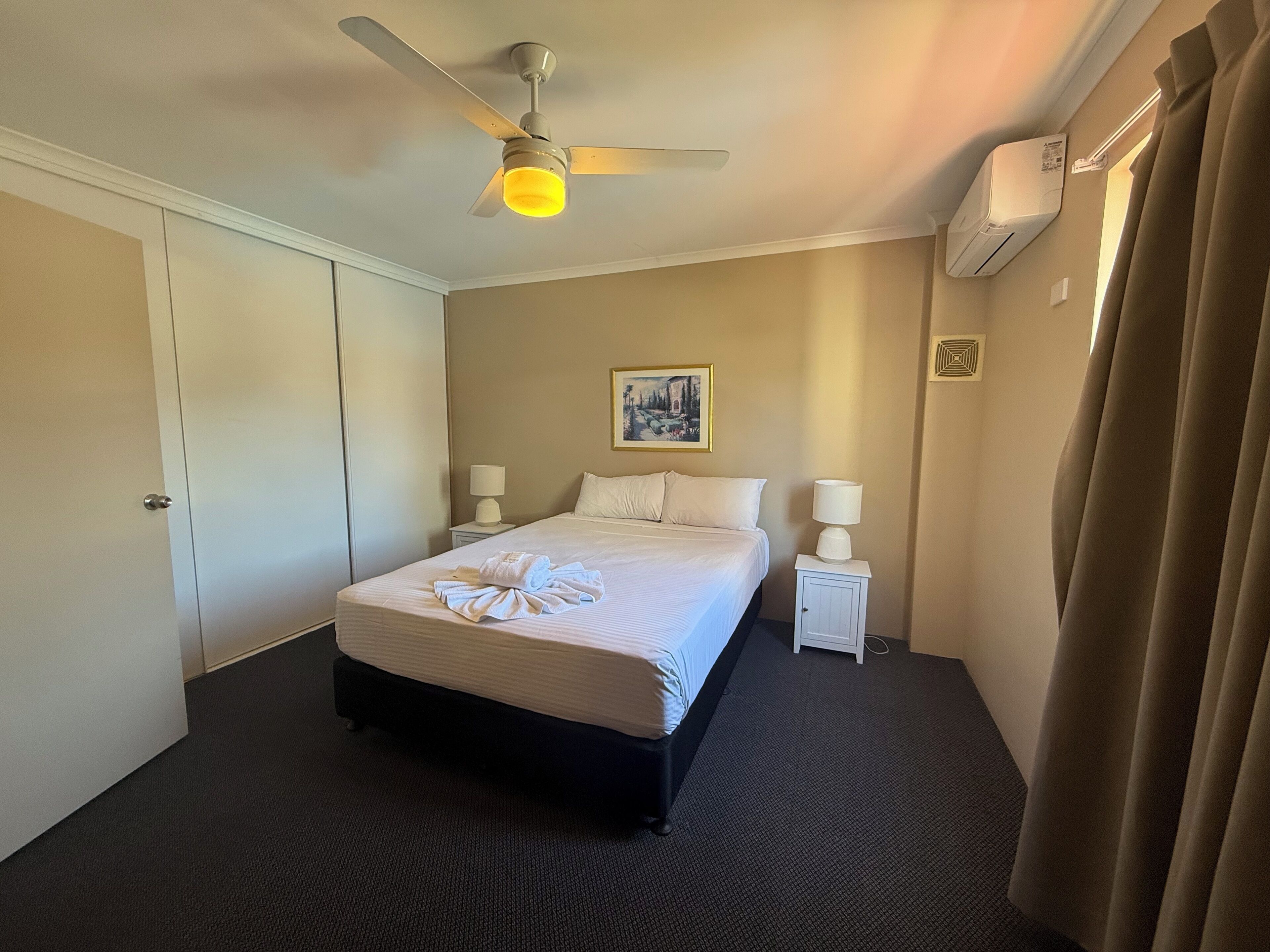 A hotel room with a bed, two bedside tables, a ceiling fan, an air conditioning unit, and a painting on the wall.