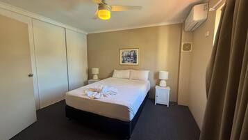A hotel room with a bed, two bedside tables, a ceiling fan, an air conditioning unit, and a painting on the wall.