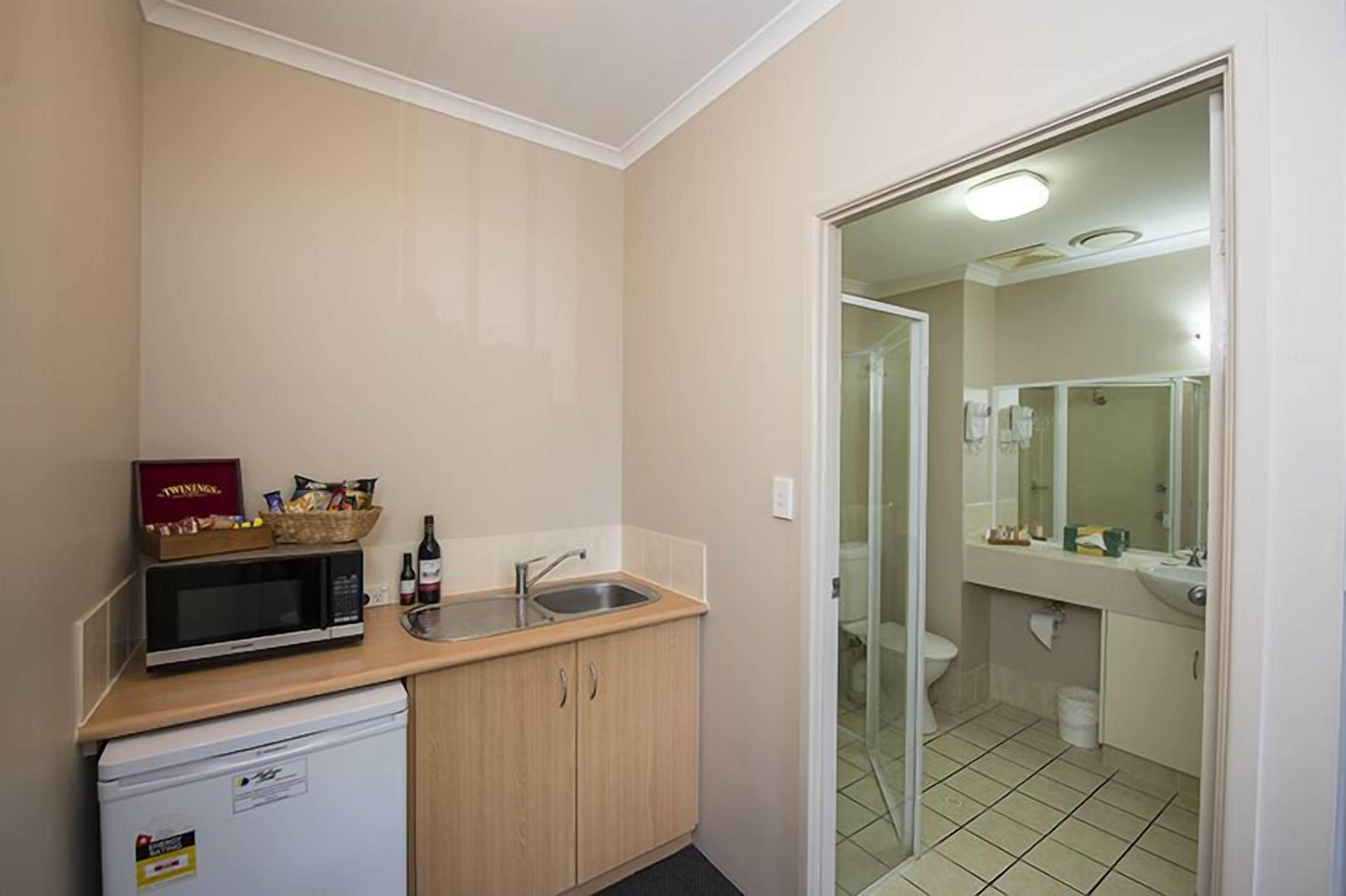 A compact kitchen with a microwave, sink, and a small refrigerator. A basket of snacks and a bottle of wine are on the counter.