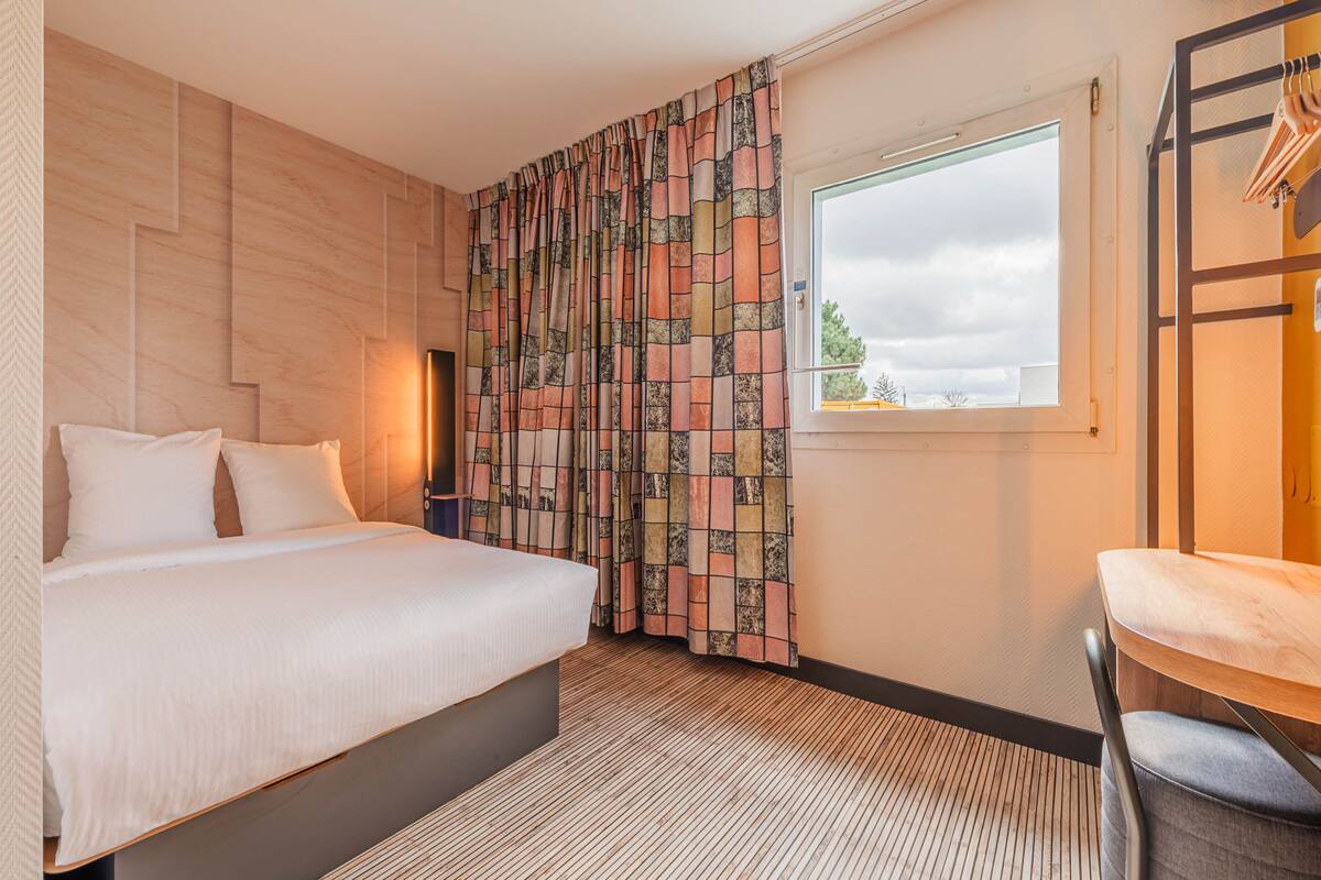A hotel room with a bed, a window with curtains, a chair, and a wall with a geometric pattern.