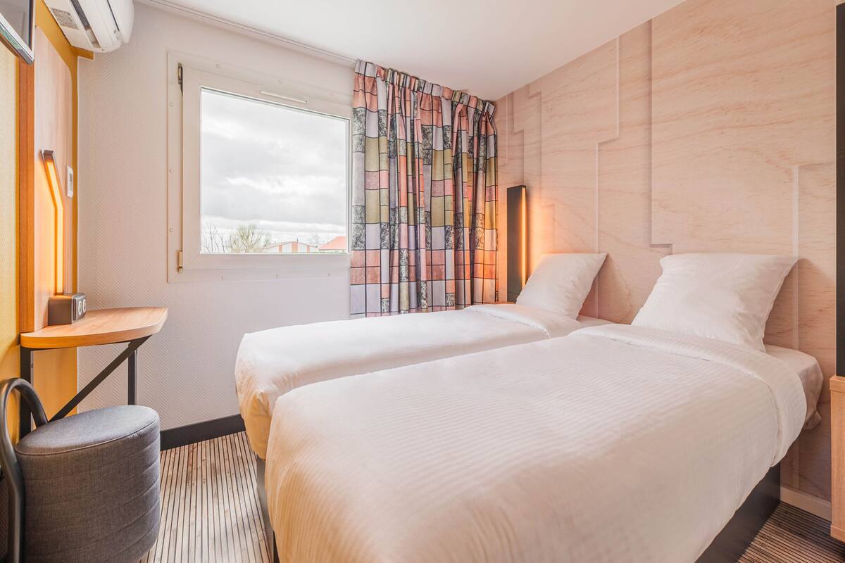 A hotel room with two beds, a small table, a chair, and a window with a view.