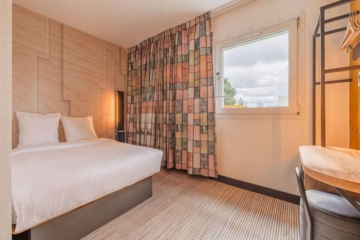 A hotel room with a bed, a window, a chair, and a wall with a geometric pattern.
