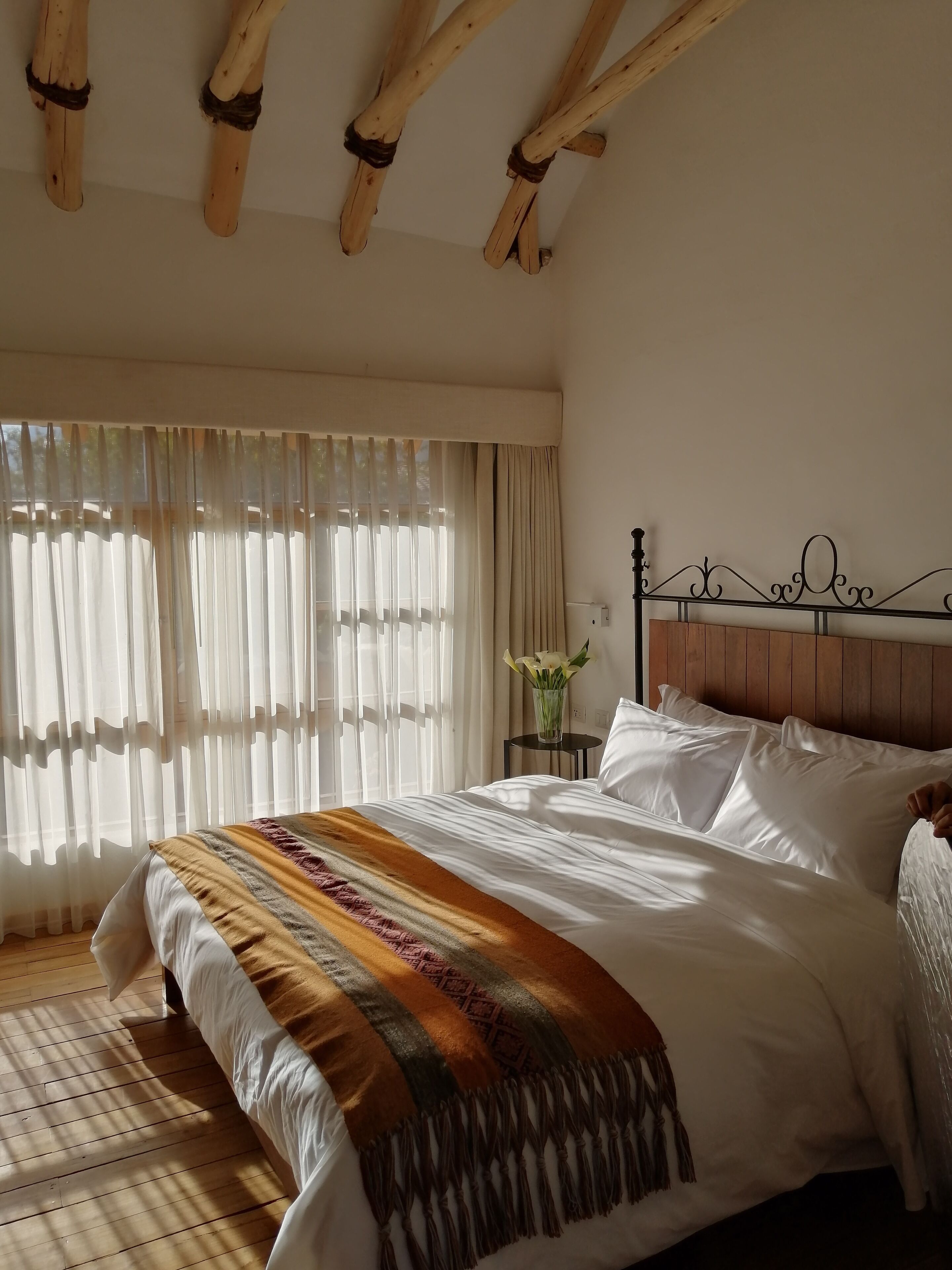 A bedroom with a wooden ceiling, a bed with white linens, a decorative throw blanket, and a nightstand with a vase of flowers.