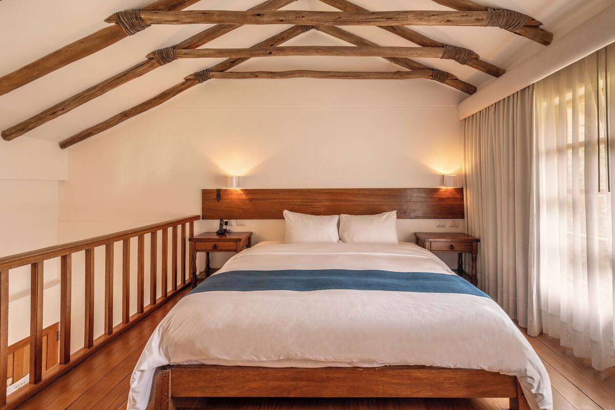 Une chambre avec un plafond en bois, un grand lit avec une literie blanche et bleue, deux tables de chevet et une petite table de nuit.