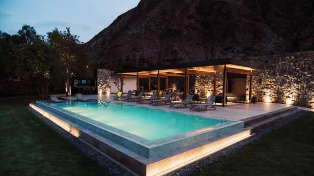 A modern outdoor pool area with lounge chairs and a covered seating area, surrounded by a stone wall and a mountain backdrop. - La Casona de Yucay Hotel (Yucay)