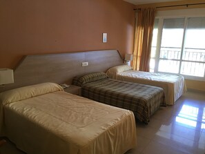 Quarto de hotel com duas camas, cabeceira, janela com cortinas e uma luminária fixada na parede.