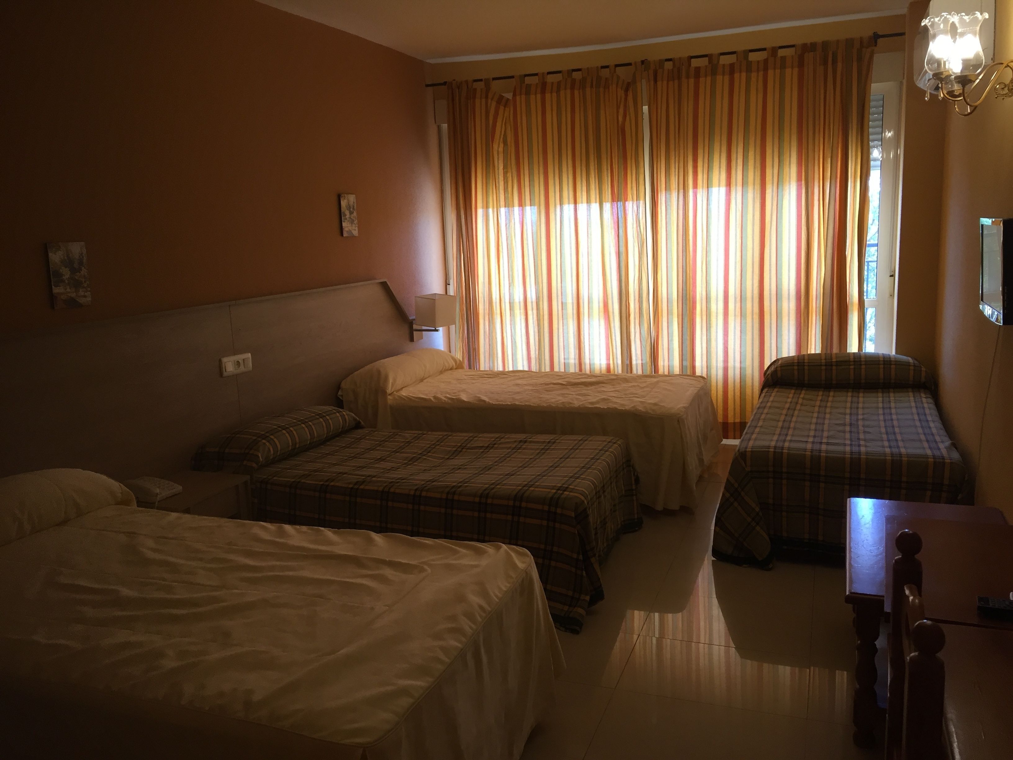 Habitación de hotel con tres camas, una mesita y un ventanal con cortinas.