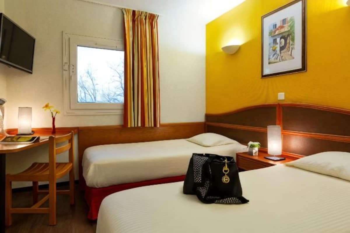 Habitación de hotel con dos camas, un escritorio con silla, televisor y ventana con cortinas.