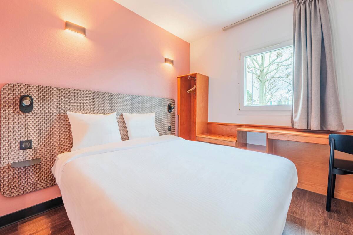 A modern hotel room with a large bed, a wooden desk, and a window with a view of trees.