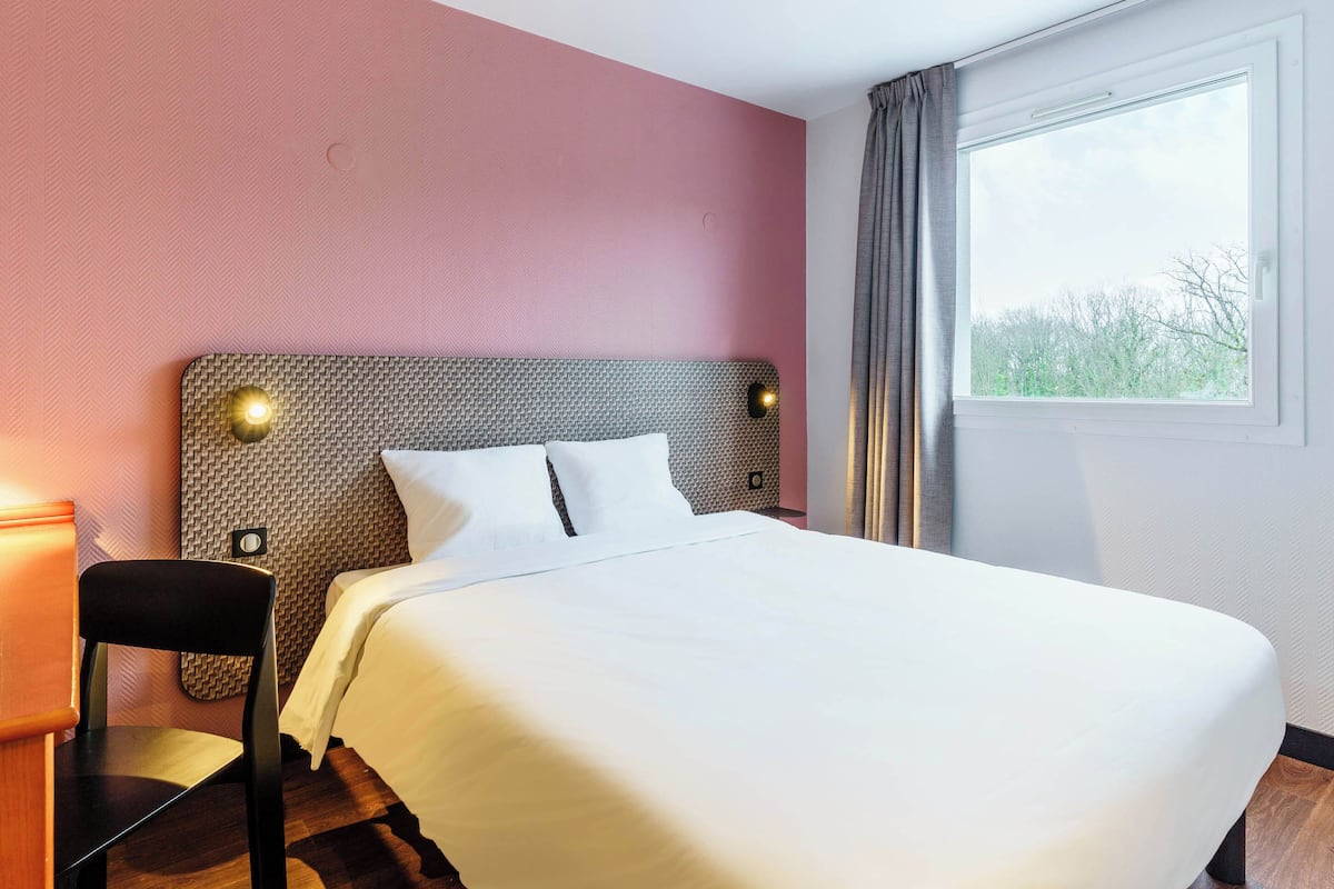 Une chambre d’hôtel avec un grand lit, une chaise, une table de chevet et une fenêtre avec des rideaux.