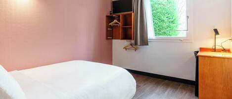 A hotel room with a bed, a wooden desk, a hanging coat rack, a window with a view of greenery, and a wall-mounted TV.