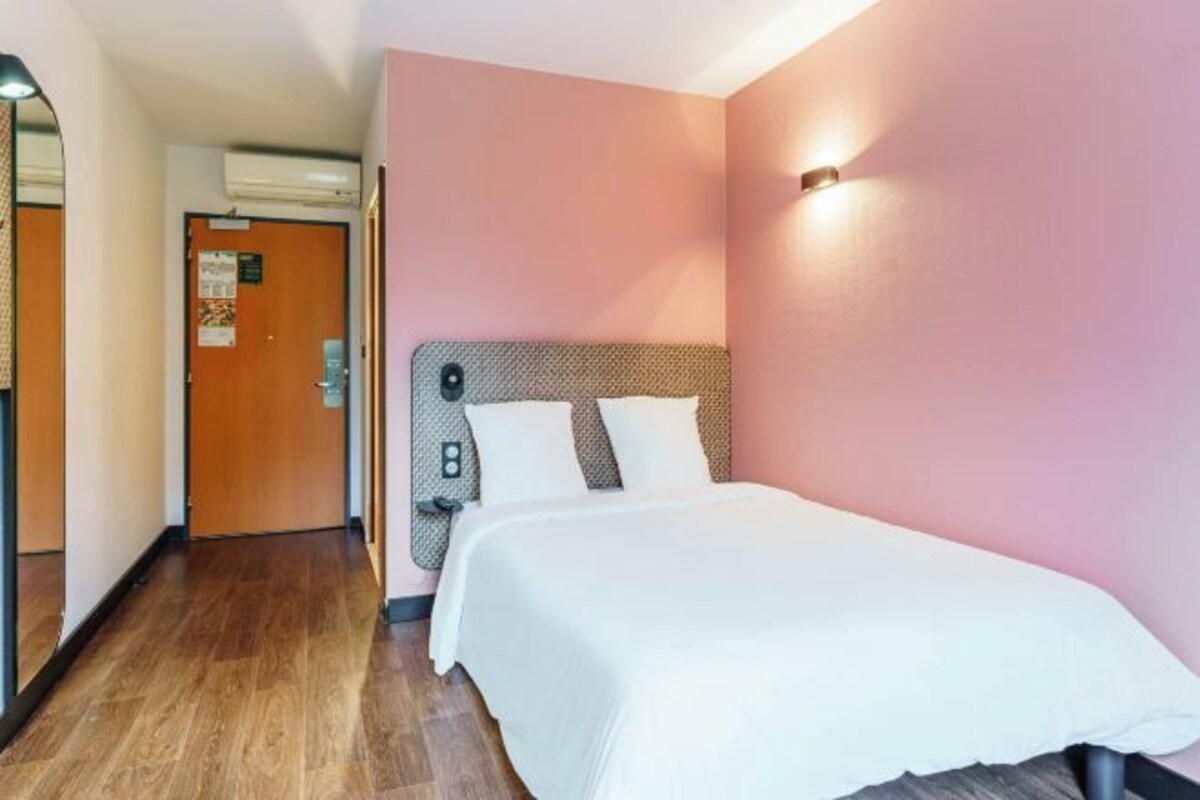A hotel room with a bed, a wooden floor, a pink wall, and a door with a bulletin board.