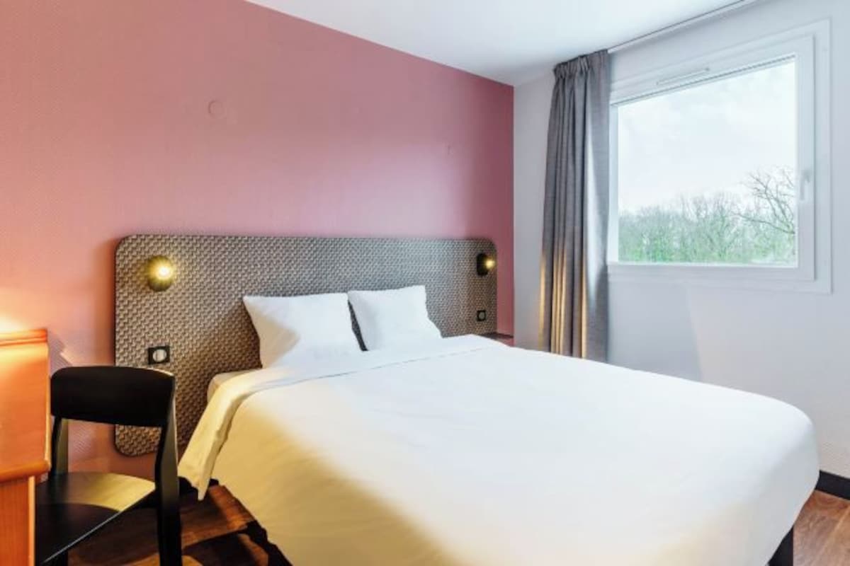 A hotel room with a large bed, a chair, a headboard with wall-mounted lights, and a window with curtains.