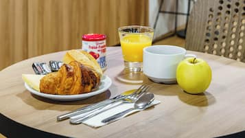 A breakfast setting with croissants, a glass of orange juice, a white cup, a yellow apple, and a container of yogurt on a round table.