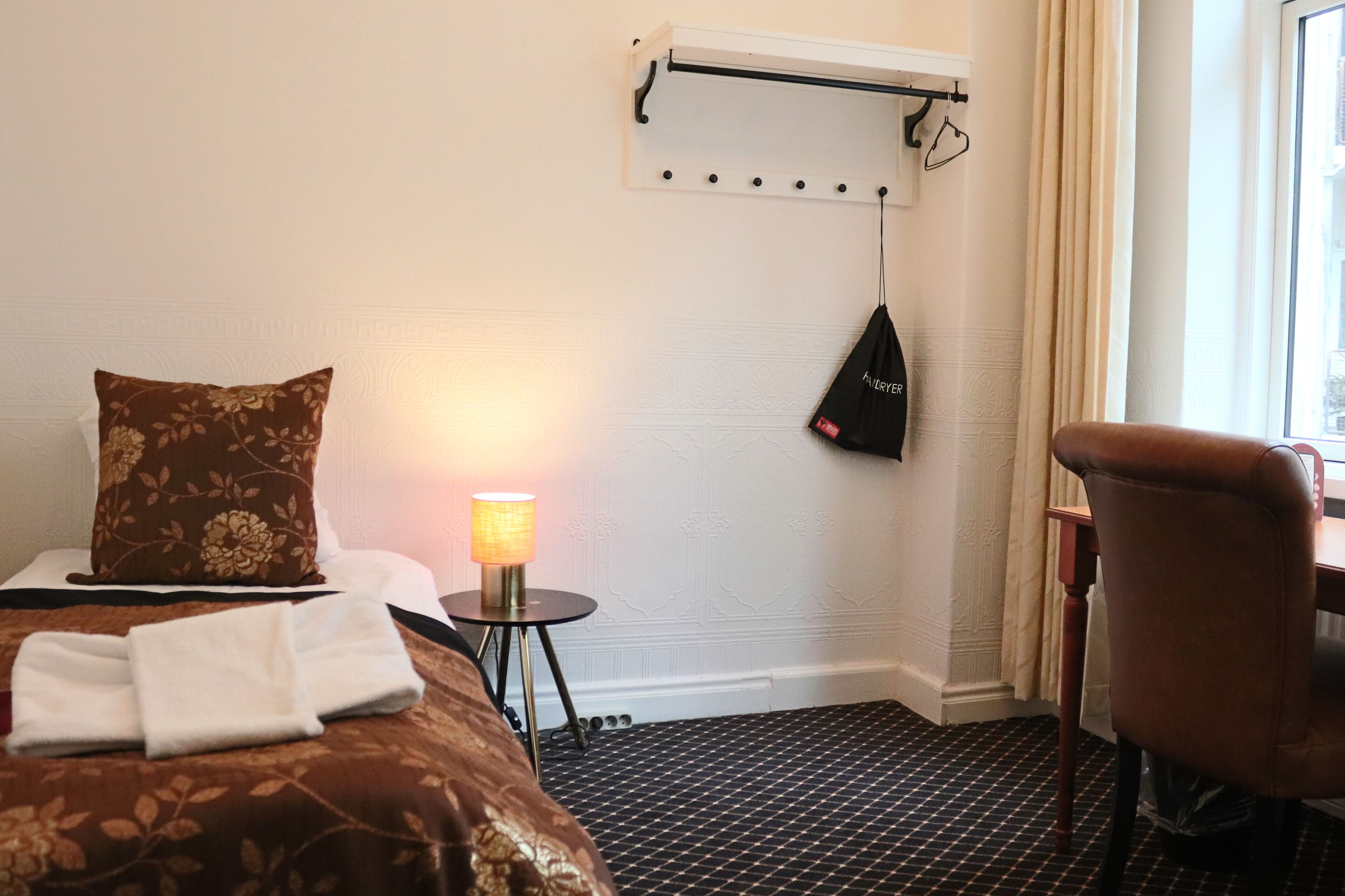 A hotel room with a bed, a bedside table with a lamp, a chair, and a window with curtains.