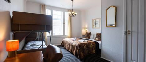 Una habitación de hotel con literas, un escritorio con lámpara, una cama con colcha estampada, un candelabro y una puerta con espejo.