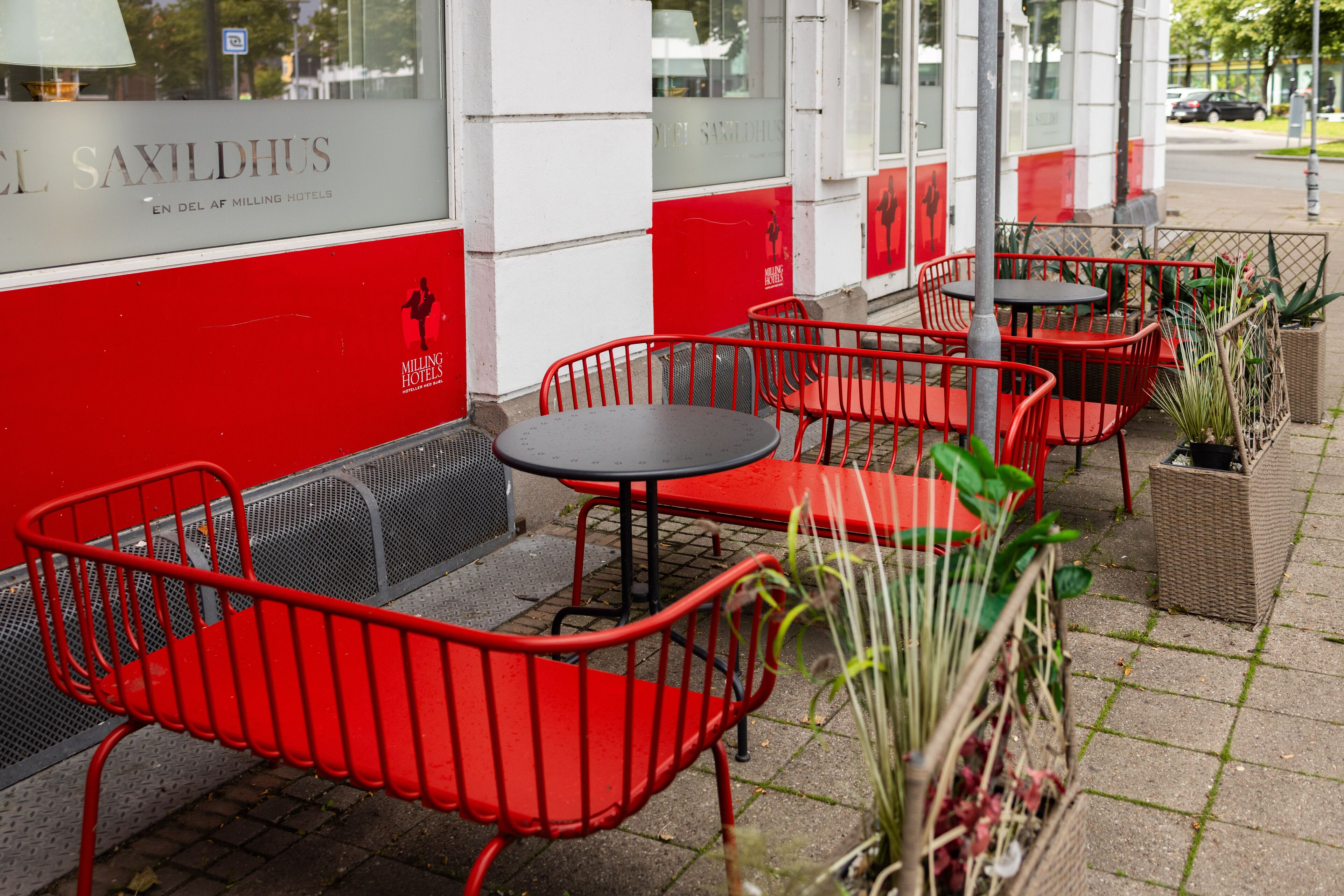 A hotel exterior with red metal chairs and tables, a tiled pavement, and potted plants.