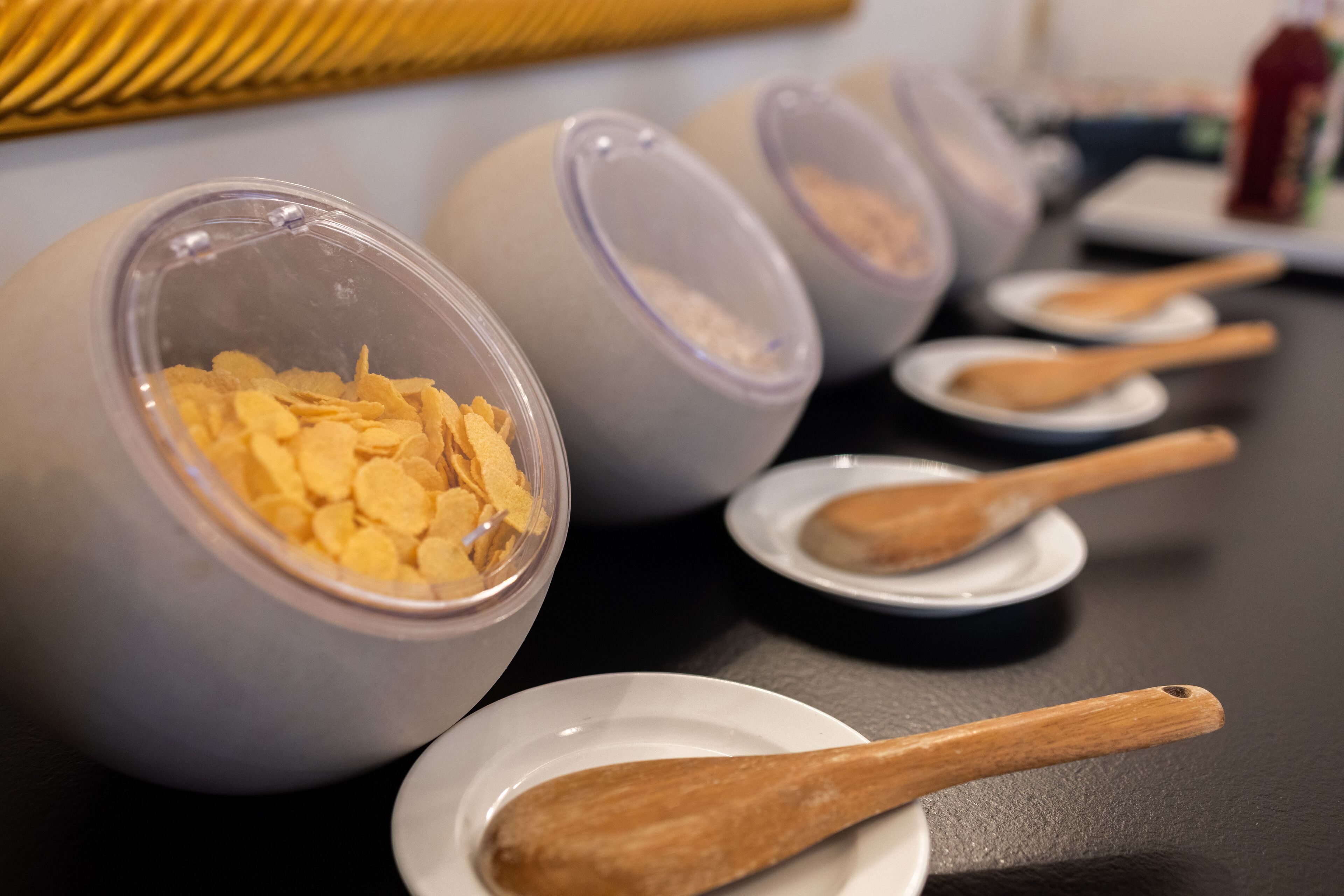 Plusieurs récipients transparents contenant des céréales sont posés sur une table, accompagnés de cuillères en bois et de petites assiettes blanches.