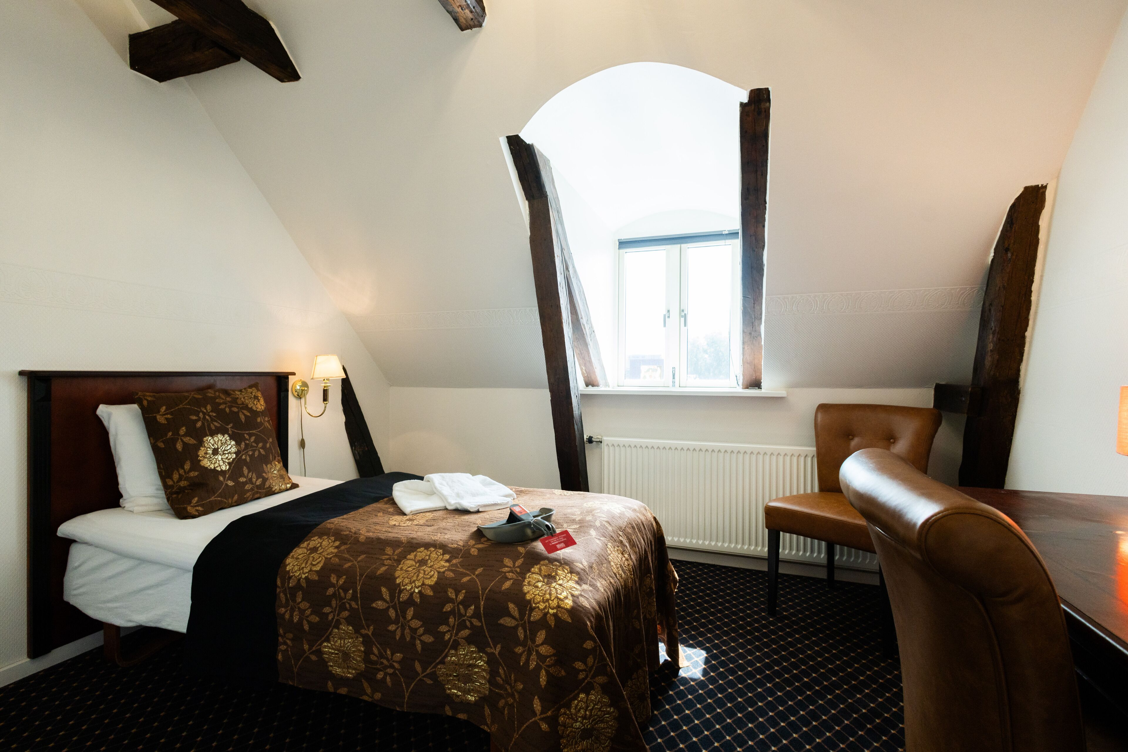 A hotel room with a bed, a chair, a desk, a window, and a radiator.