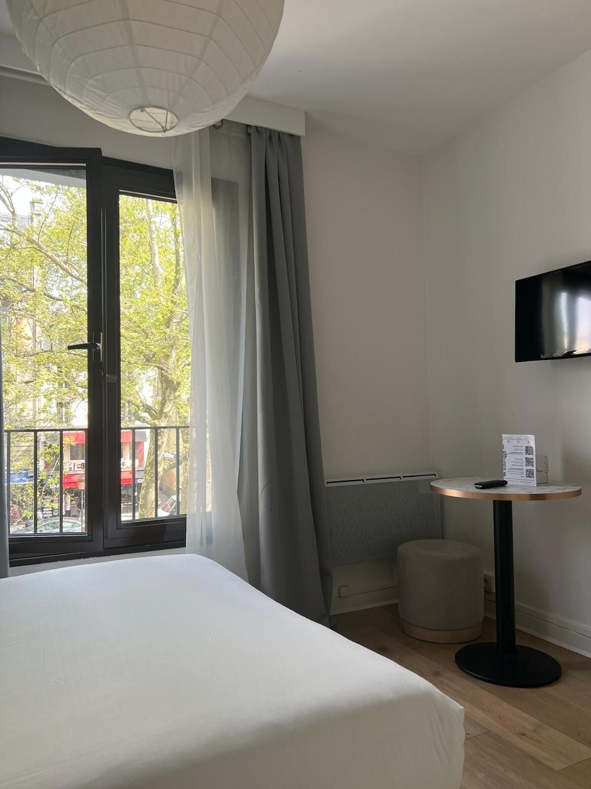 Habitación de hotel con cama, una mesita con control remoto, televisor montado en la pared y ventana con cortinas.