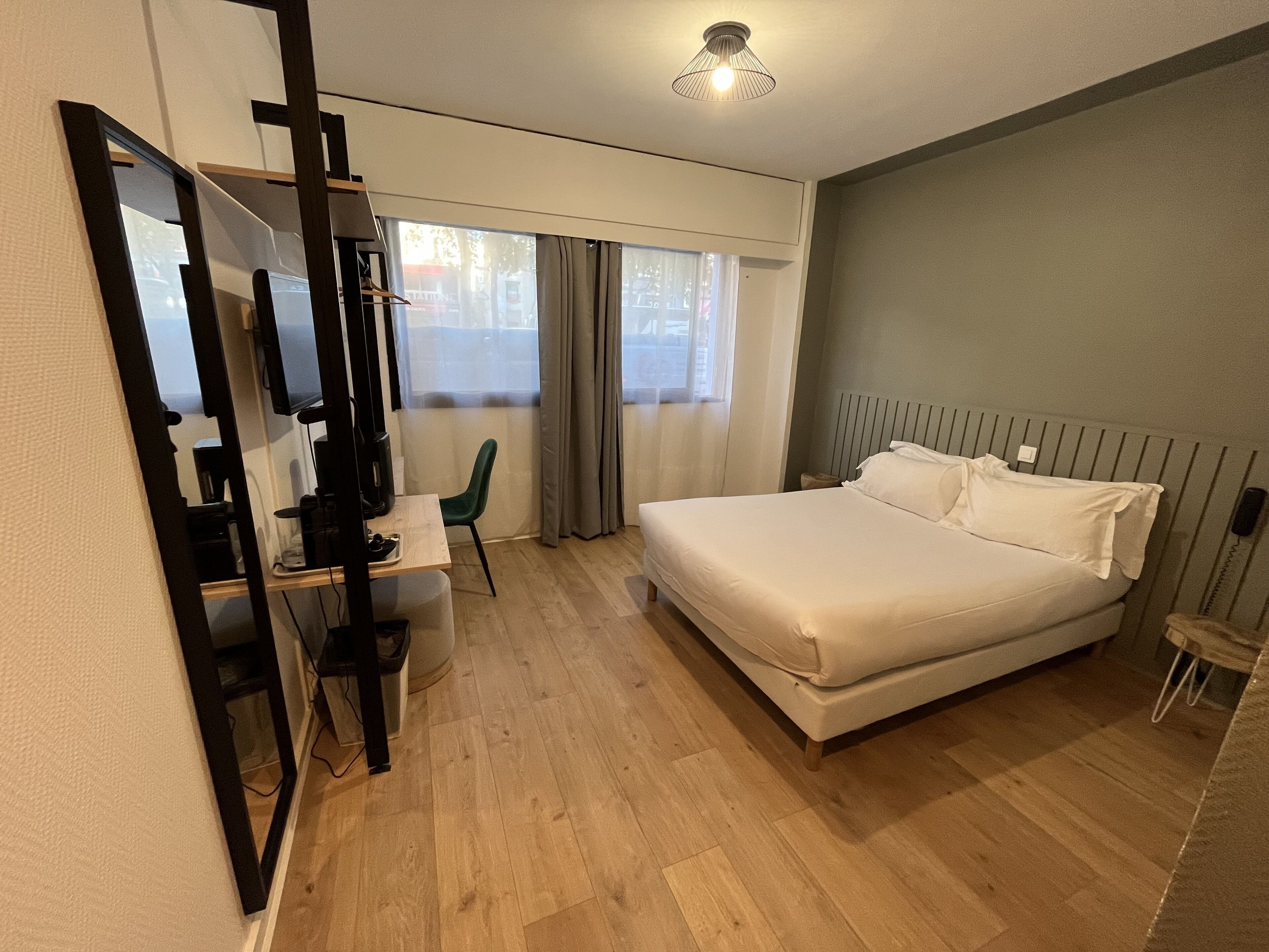 A modern hotel room with a large bed, a desk with a chair, a television, and a window with curtains.