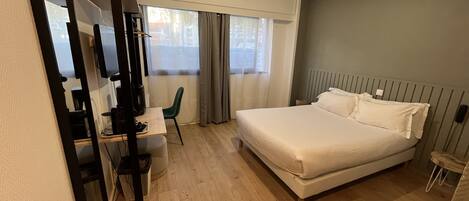A modern hotel room with a large bed, a desk with a chair, a television, and a window with curtains.