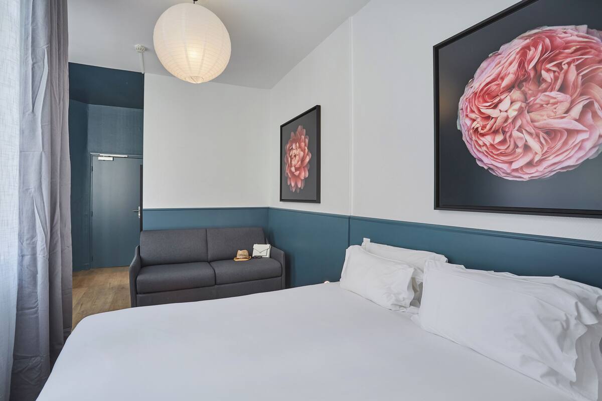 Una habitación de hotel con una cama, un sofá gris, una obra de arte floral grande en la pared y una lámpara del techo.